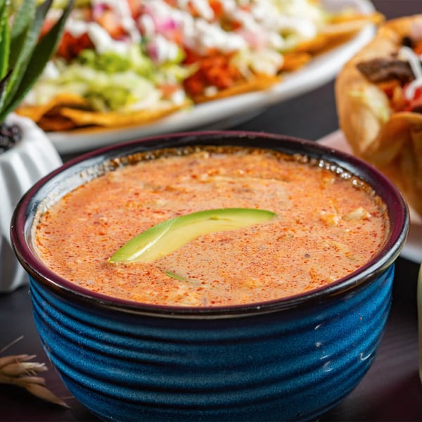 A blue bowl of creamy Mexican soup with a slice of avocado, with nachos in the background.
