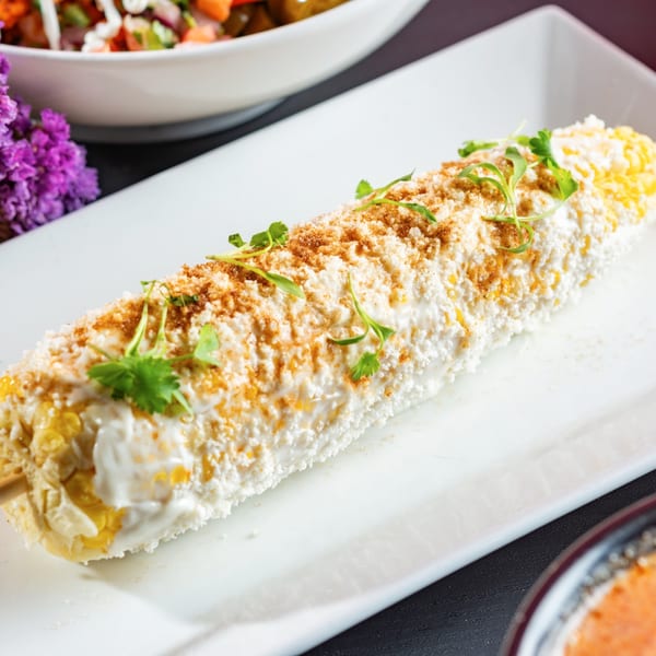 Elote, Mexican street corn, on a white plate, topped with cotija cheese, chili powder, and cilantro.