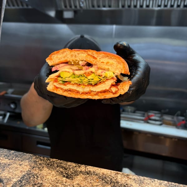 A chef in black gloves holds a large, fresh Mexican torta sandwich filled with chicken, lettuce, and onions.