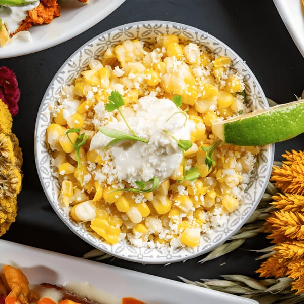 A bowl of Mexican street corn (Esquites) with cotija cheese, crema, cilantro, and a lime wedge.